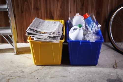 Two plastic recycling bins placed on a concrete floor in an indoor or garage setting. The yellow bin on the left contains neatly stacked newspapers and magazines. The blue bin on the right holds clear plastic bottles and containers, some with caps, and a few empty water bottles. Behind the bins, a wooden wall with a vertical panel design is visible. To the left, part of a metal shelving unit can be seen, and on the right, a portion of a bicycle wheel is partially in view. The scene suggests an area used for household waste or recycling collection, consistent with a rubbish removal service environment in the Kensington area. The lighting appears to be natural or from ambient indoor sources, casting soft shadows on the floor and highlighting the textures of the plastic, paper, and wood surfaces.