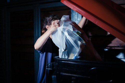 A person disposing of transparent plastic bags filled with assorted waste materials into a large, black skip positioned outdoors, with a wooden structure and blue metal framework visible in the background. The individual is partially obscured, holding an open bag over the skip as they unload rubbish. The scene is illuminated by natural light, suggesting daytime, and reflects a typical rubbish clearance activity on a residential or commercial property in London, near Kensington. The setting indicates an organized effort by Kensington House Clearance to remove waste from a property, with the waste being primarily plastic and household rubbish typical of clearance services in the area.