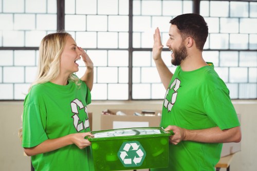 A man and a woman wearing bright green recycling T-shirts are indoors against a backdrop of large window panes divided by dark metal frames, suggesting an industrial or warehouse setting. The woman, on the left, has blonde hair and is smiling with her hand raised in a gesture of greeting or agreement. The man, on the right, has dark hair and a beard, and is also smiling while giving a high-five or engaging in friendly communication. Between them, they hold a green recycling bin with a white recycling symbol on the front, indicating their involvement in waste or rubbish collection. The scene reflects an environmentally conscious activity, possibly part of a rubbish removal service or community recycling effort. The background includes a glimpse of cardboard boxes and a concrete or tiled floor, emphasizing the indoor, operational context relevant to waste management services offered by companies such as Kensington House Clearance in London postal code area, supporting local rubbish clearance tasks.
