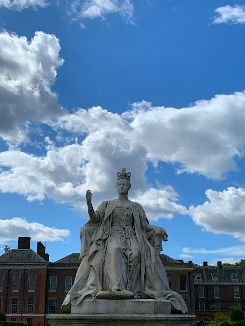A stone statue of a regal female figure wearing a crown and draped in flowing robes, seated on a pedestal with her right arm raised and her left hand resting on her lap, located outdoors against a bac